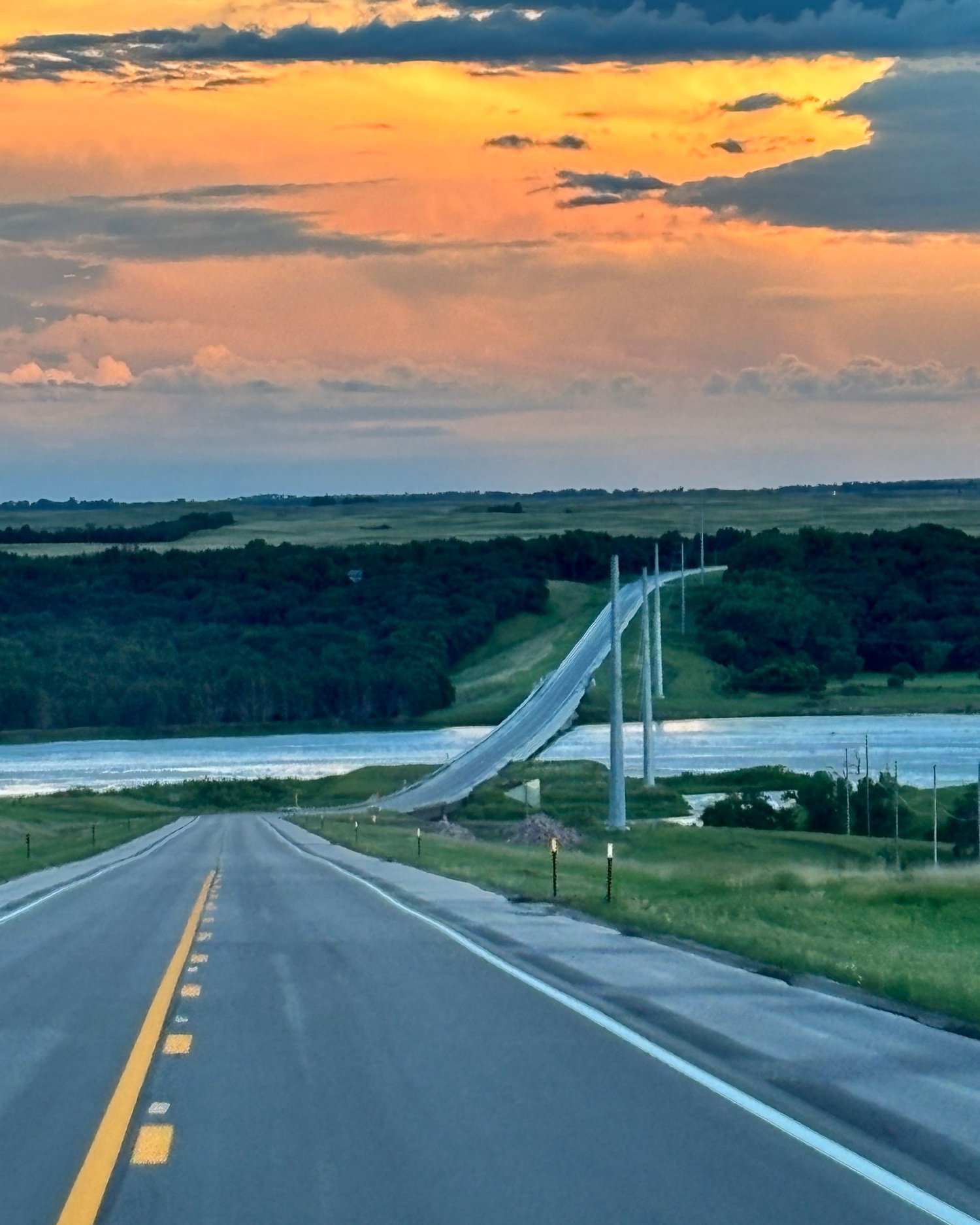 Scenic view of a highway leading towards a bridge over water, with a vibrant sunset sky featuring orange and blue hues, surrounded by lush green landscapes and rolling hills.