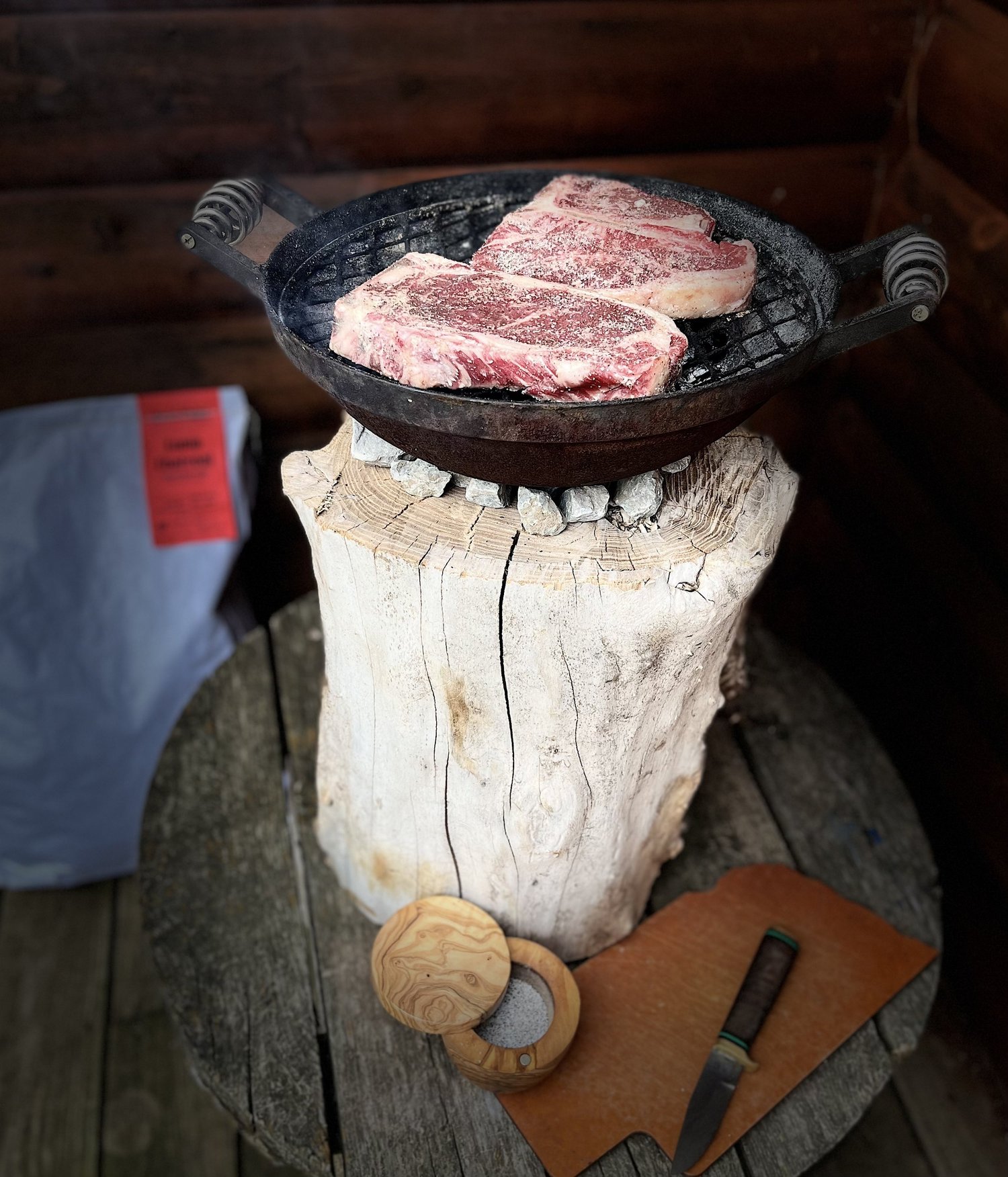 Grilling two marbled ribeye steaks in a cast iron skillet over a rustic wooden stump, with a wooden bowl of salt and a knife on a leather cutting board nearby.