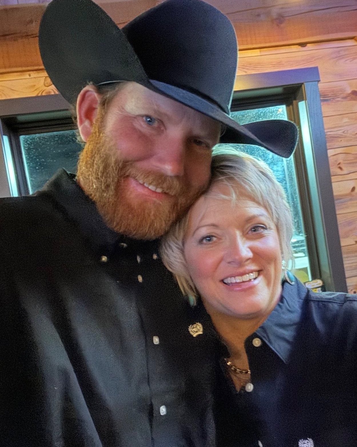 A smiling couple poses closely together, the man wearing a large black cowboy hat and a dark shirt with buttons, while the woman has short blonde hair and a matching dark shirt. The background features a wooden interior and a window, suggesting a cozy setting.